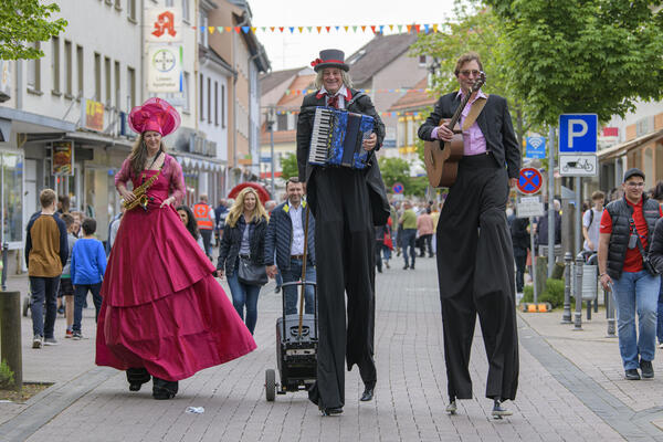 Drei Stelzenl&auml;ufer flanieren mit ihren Musikinstrumenten durch die Innenstadt.