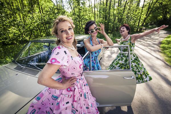 In ihren bunten Petticoatkleidern posiert das Musik-Trio "The Wonderfolleins" vor einem Oldhimer in der Natur.