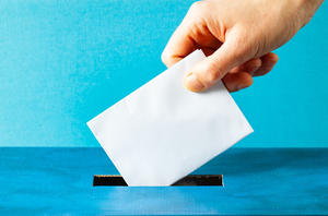 Eine Hand steckt ein Papier in eine Wahlurne.