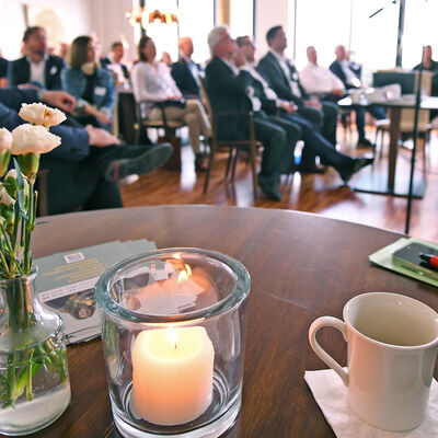 Im Vordergrund ein Tisch mit Kerze, Blumen und Kaffeetasse, w&auml;hrend im Hintergrund die G&auml;ste beim Unternehmerfr&uuml;hst&uuml;ck dem Vortrag lauschen.