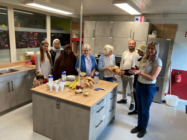 Acht aufgereihte Personen vor einer gedeckten K&uuml;cheninsel f&uuml;r ein gemeinsames Fr&uuml;hst&uuml;ck im Rahmen des Begegnungsprojekts &raquo;Kaffee und Kakao&laquo;.