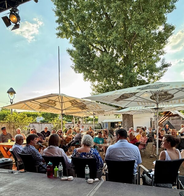 Blick von der B&uuml;hne aus auf die Teilnehmenden und das Publikum im Biergarten im Rahmen einer Podiumsdiskussion.