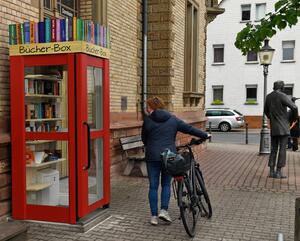 "Bücher-Box" vor dem Gebäude der alten Schule in Dornheim. Davor eine Frau, die ihr Fahrrad schiebt und im Vorbeigehen in die "Bücher-Box" schaut.